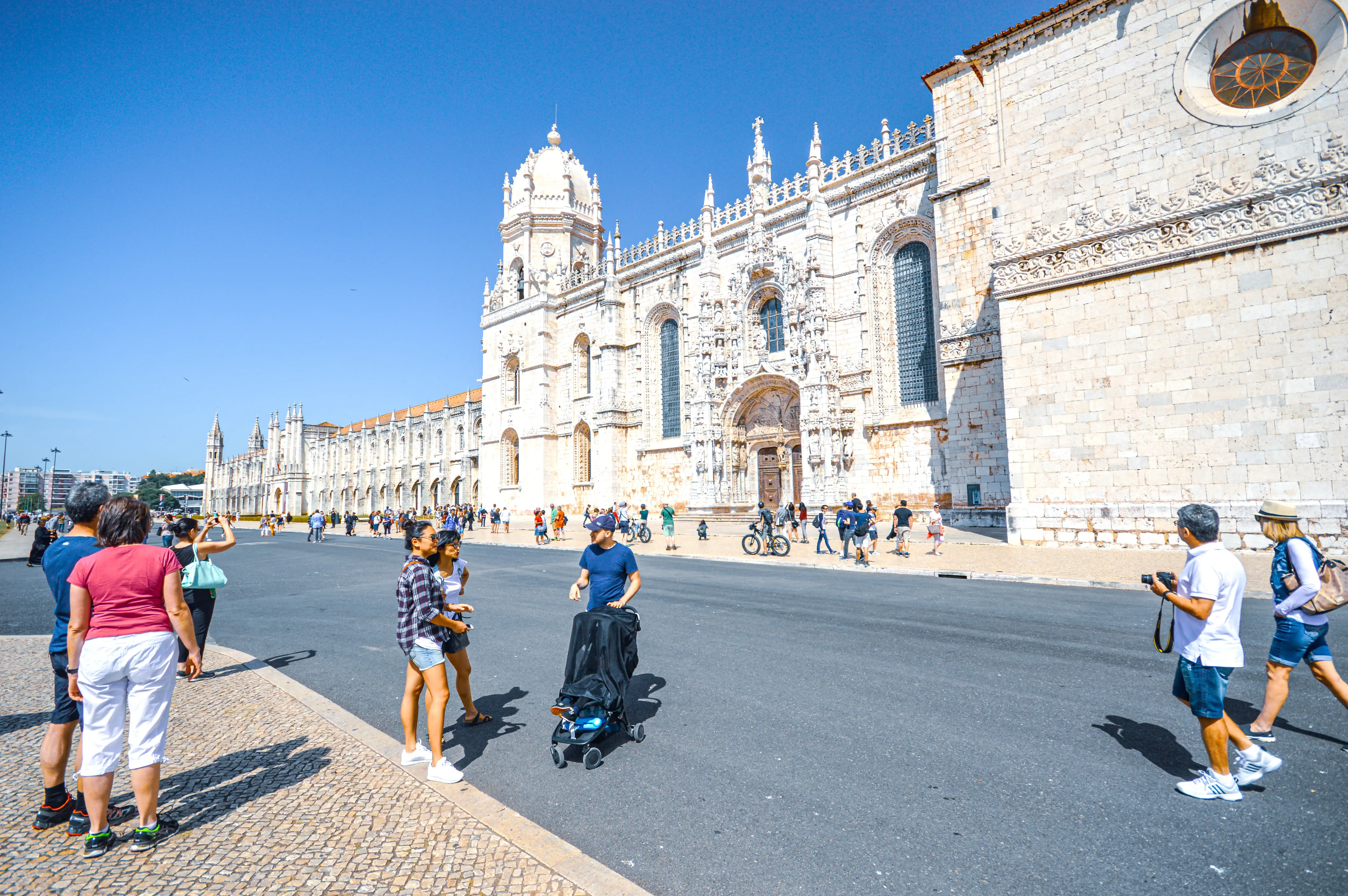 Wide view of the Jerónimos Monastery in Lisbon, Portugal, under a clear blue sky, with numerous tourists walking on the pavement and road in front of the historic building.